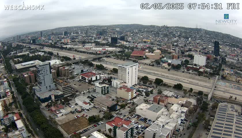 An aerial view of a city on an overcast day shows a wide roadway cutting through a densely built urban area, with various buildings and parking lots visible.