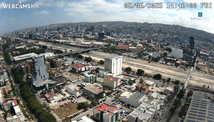 An aerial view of a city on a partly sunny day shows a wide, dry riverbed cutting through the urban landscape, with numerous buildings and roads visible.