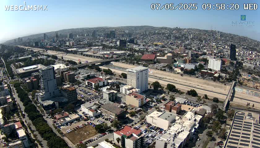 An aerial view on a sunny day shows a sprawling city with a long, wide roadway running through it, alongside a river or dry riverbed.