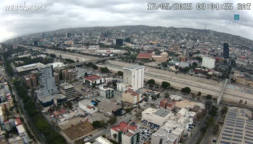An aerial view of a city under a cloudy sky, showing a wide roadway cutting through a densely populated urban area with numerous buildings and sparse vegetation.