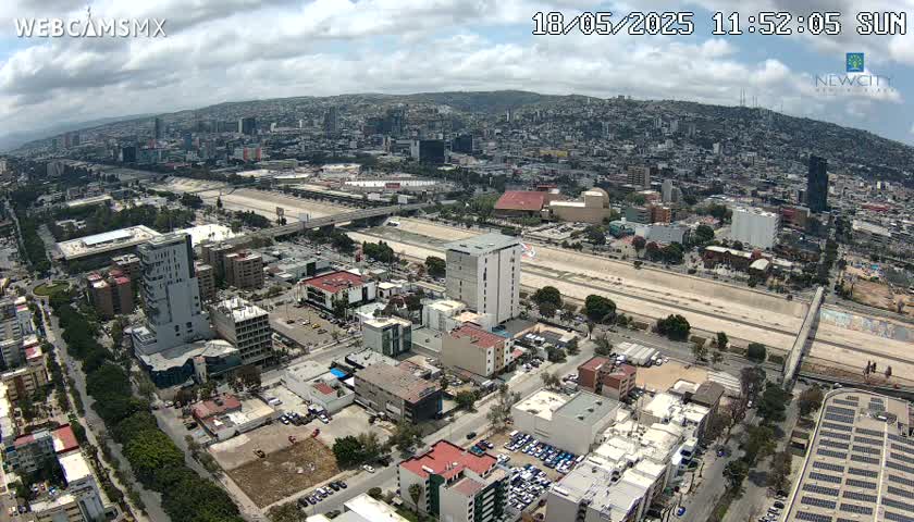 An aerial view of a city on a partly cloudy day, showing a large highway cutting through an urban landscape with many buildings and parking lots.