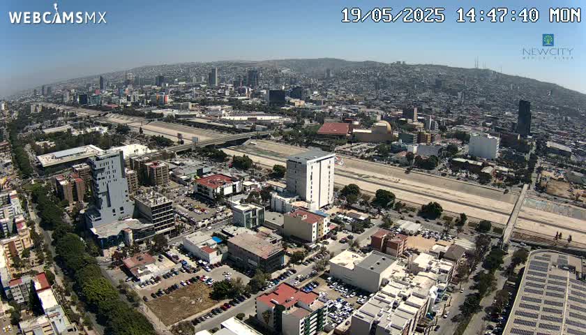 An aerial view of a sprawling city on a sunny day, showing a wide roadway and a riverbed running through a densely populated area.