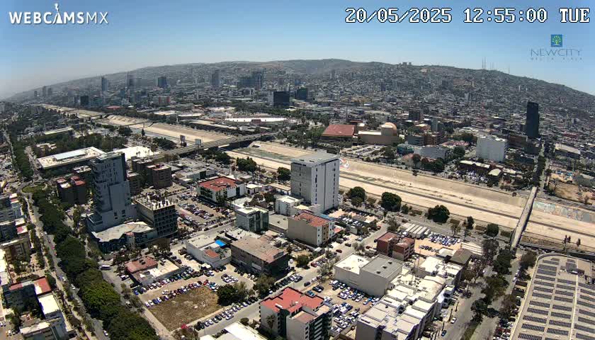 An aerial view shows a sprawling city under a clear, sunny sky, with a wide roadway and a river or canal running through it.