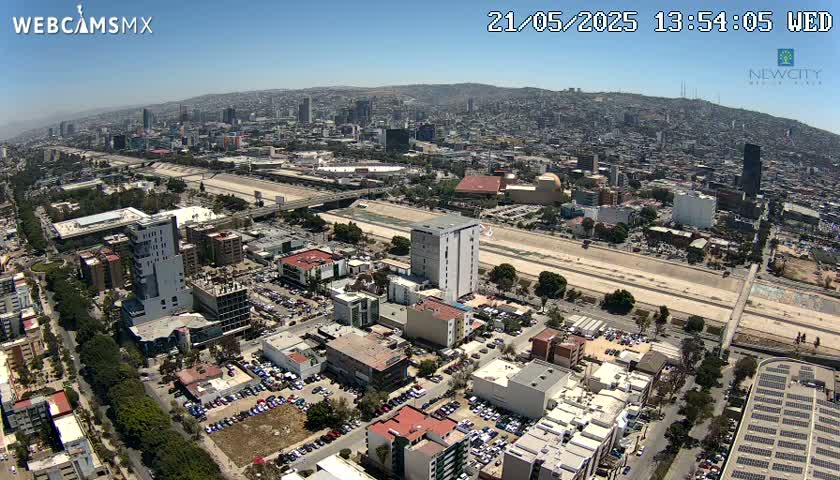 An aerial view shows a sprawling city on a sunny day, with a wide, dry riverbed cutting through a section of the urban landscape.