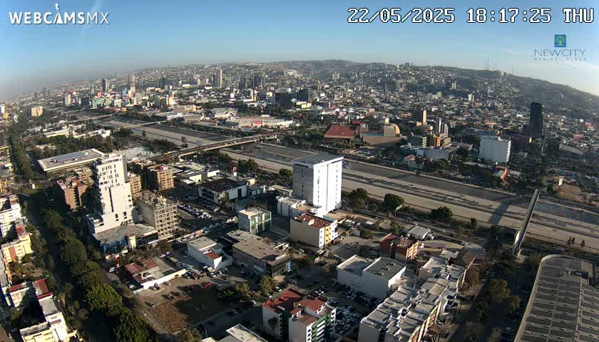 An aerial view of a city on a sunny day, showing a wide highway cutting through a sprawling urban landscape.