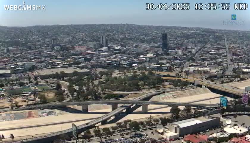 An aerial view shows a sprawling city on a clear, sunny day, with a complex network of highways and overpasses intersecting near a large park-like area.