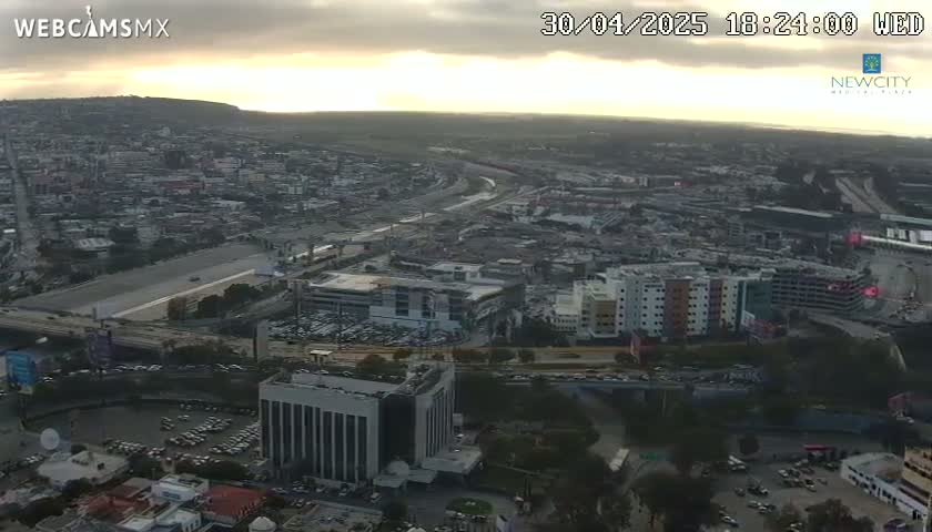 An aerial view of a city at sunset, showing highways, buildings, and parking lots under a cloudy sky.