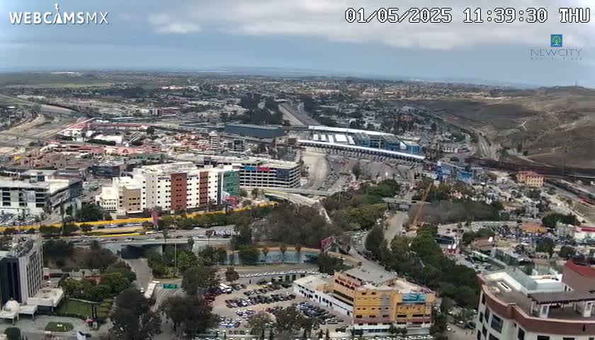 An aerial view shows a partly cloudy day over a city with numerous buildings, roads, and a large parking lot near a border crossing.