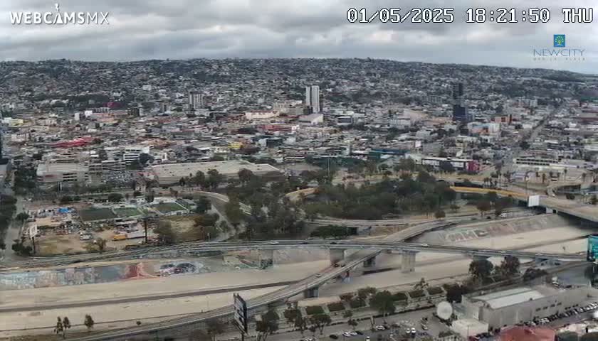 An expansive view of a city under a cloudy sky shows a sprawling cityscape with numerous buildings, roads, and a large overpass system.