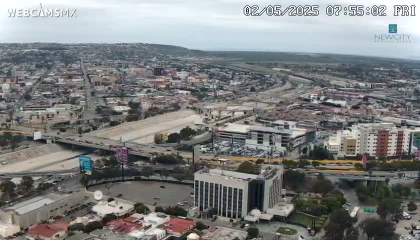 An aerial view shows a city with overcast skies, featuring a large highway system weaving through numerous buildings and parking lots.