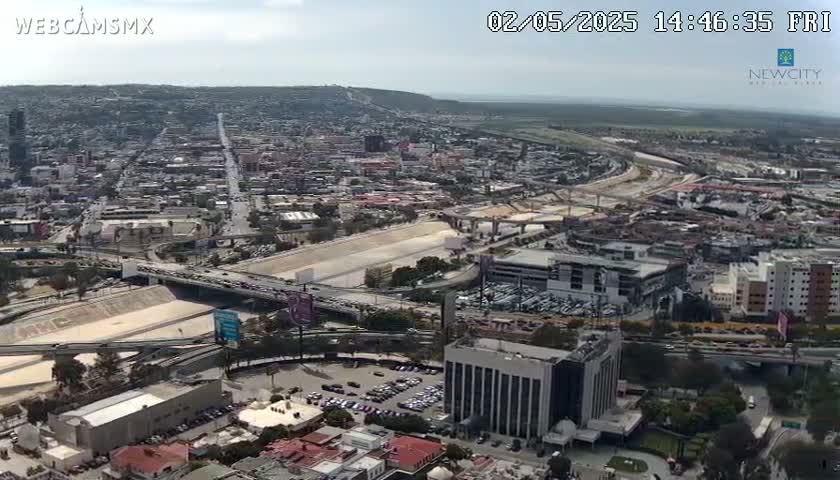 An aerial view of a city on a partly cloudy day shows a large highway system and numerous buildings, with a body of water visible in the distance.
