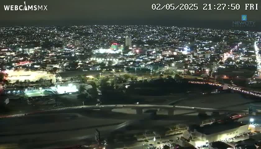 A nighttime, high-angle view of a sprawling city illuminated by countless lights, featuring a large stadium and a highway overpass.