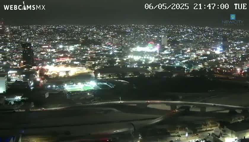 A nighttime, aerial view of a brightly lit city sprawling across a landscape, with a highway overpass visible in the foreground.