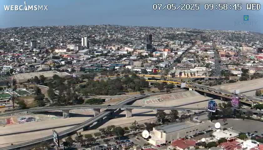 An aerial view shows a sprawling city under a clear blue sky, with a complex highway system and bridges intersecting a dry riverbed.