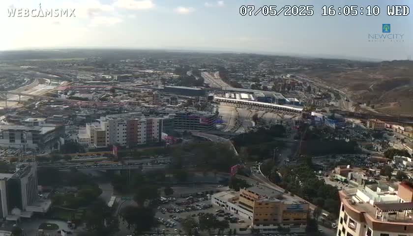 A high-angle, long shot shows a sprawling cityscape under a mostly sunny sky with a few clouds, featuring highways, buildings, and parking lots.
