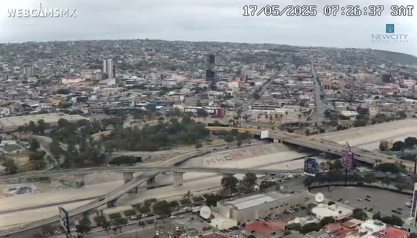 An overcast day reveals a sprawling city landscape with numerous roads and bridges intersecting, some with graffiti, alongside a dry riverbed.