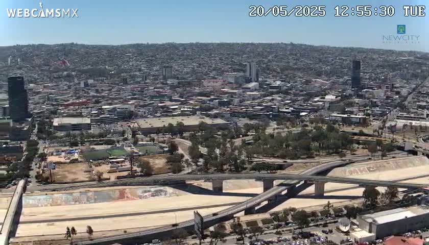 An aerial view shows a sprawling city under a clear blue sky, with several highway overpasses intersecting near a large, mural-decorated area.