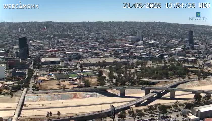 A sprawling city under a clear blue sky is viewed from above, showing a large highway system intersecting with a dry riverbed.