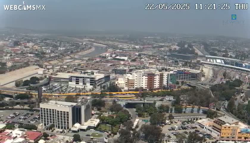 An aerial view shows a hazy city landscape with a river, roads, buildings, and parking lots on a partly cloudy day.