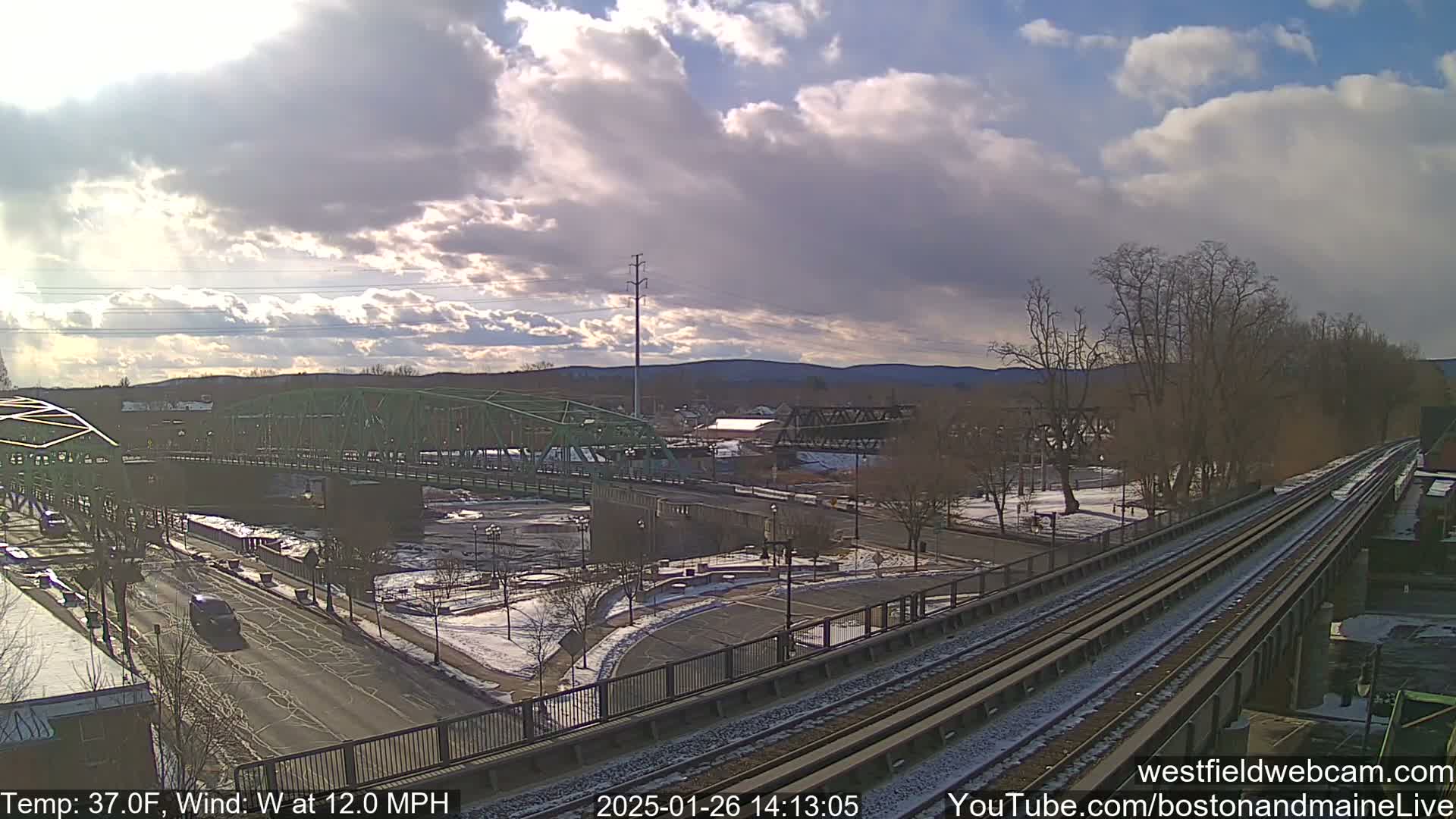 A partly cloudy day with a 12 mph wind reveals a view of a river, two bridges, train tracks, and a roadway with some light snow on the ground.