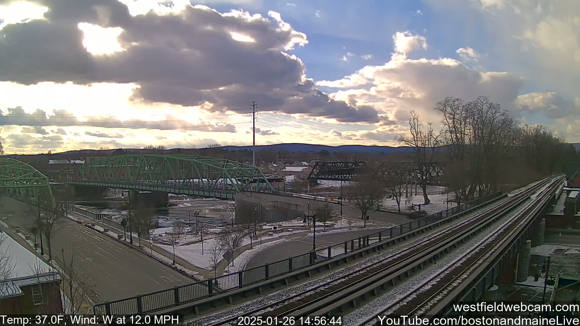 A partly cloudy sunset over a river, with two bridges and train tracks, and light snow on the ground.