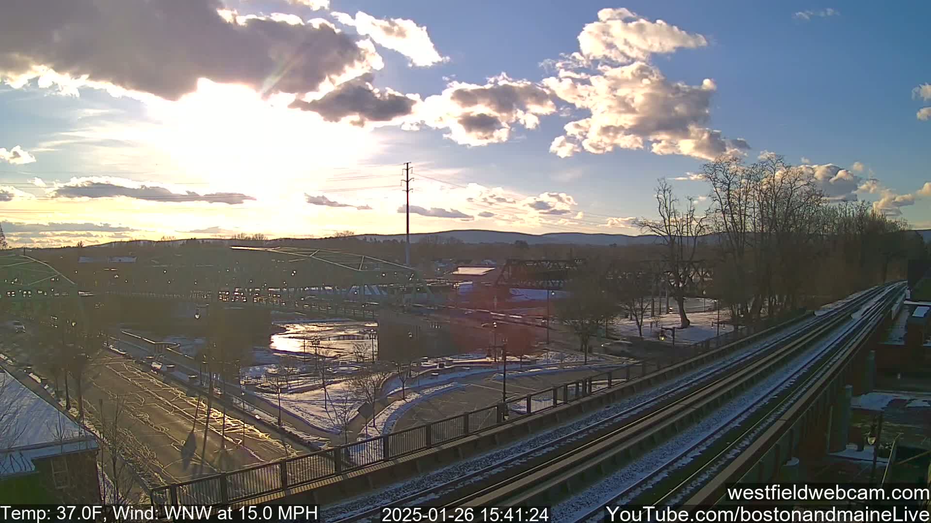 A partly sunny winter scene shows a train track overlooking a town square with some snow on the ground, under a partly cloudy sky.