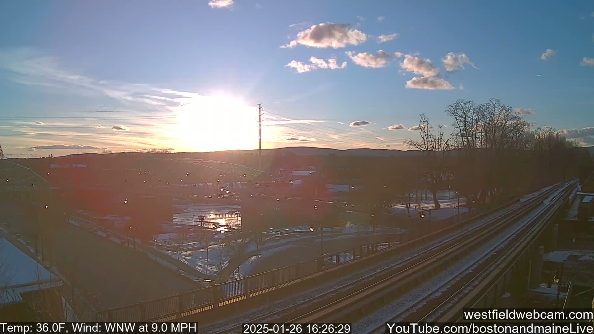Here's a description of the image: A setting sun casts a warm glow over a winter landscape of train tracks, a park, and buildings, with a light dusting of snow on the ground under a partly cloudy sky.