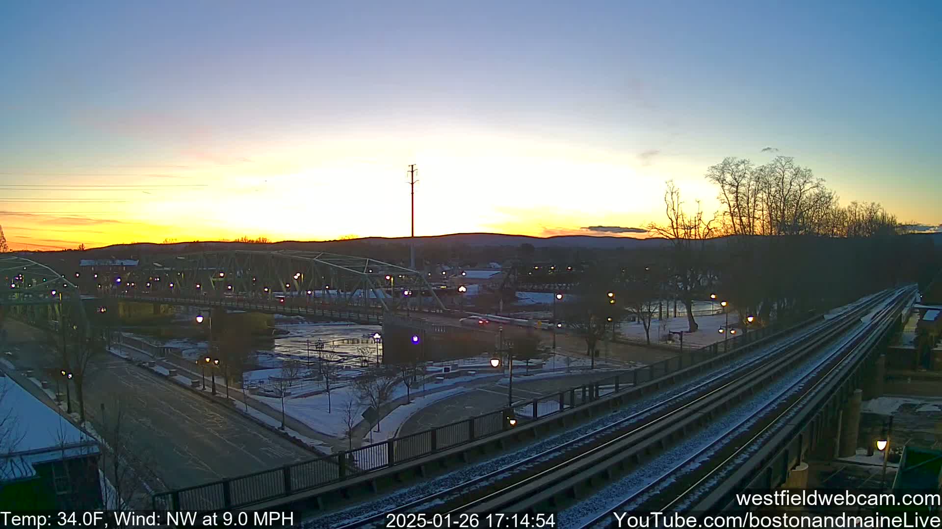 Here is a description of the image: A sunset over a snow-dusted town with a river, bridge, and train tracks, with a northwest wind at 9.0 mph and a temperature of 34 degrees Fahrenheit.
