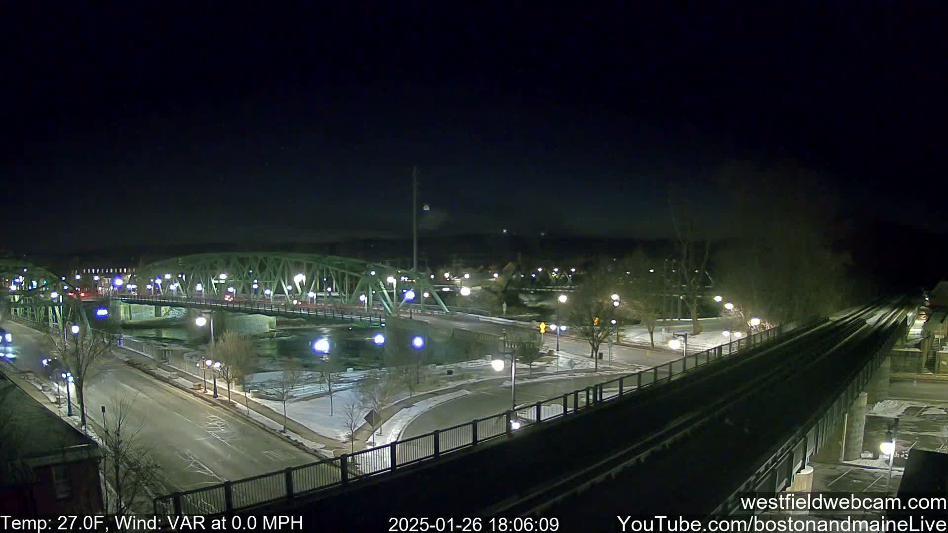 A nighttime view of a lit-up bridge spanning a river, with some snow on the ground and a temperature of 27 degrees Fahrenheit.