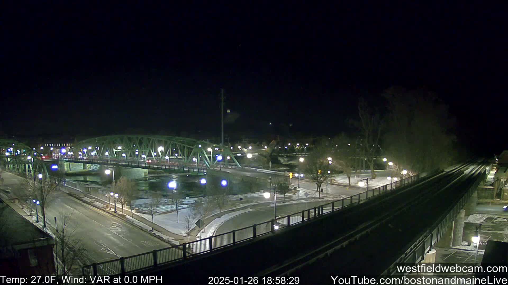 A nighttime view of a bridge over a river, with lightly snow-covered ground and a nearby train track, under a clear, cold sky.