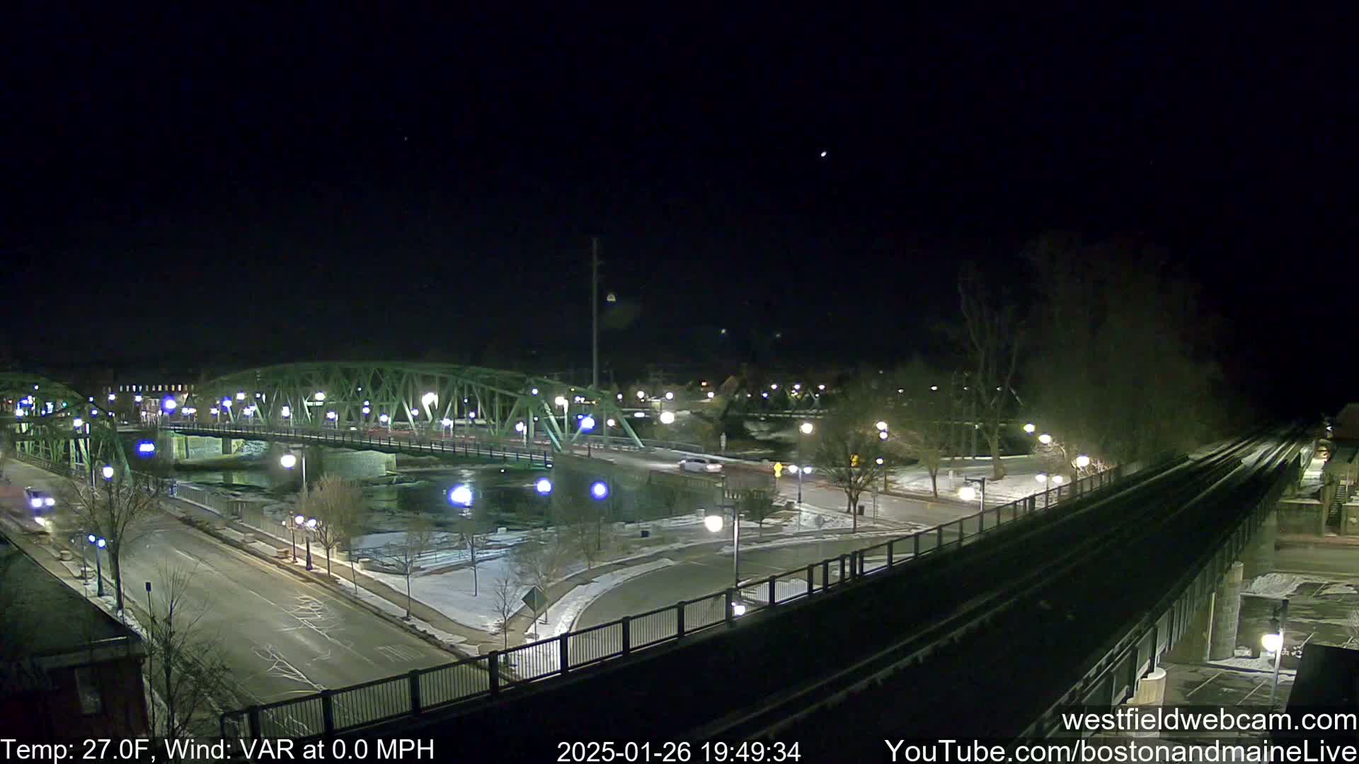 A nighttime view of a bridge over a river, with snow on the ground and a calm, clear sky, with the temperature at 27 degrees Fahrenheit.