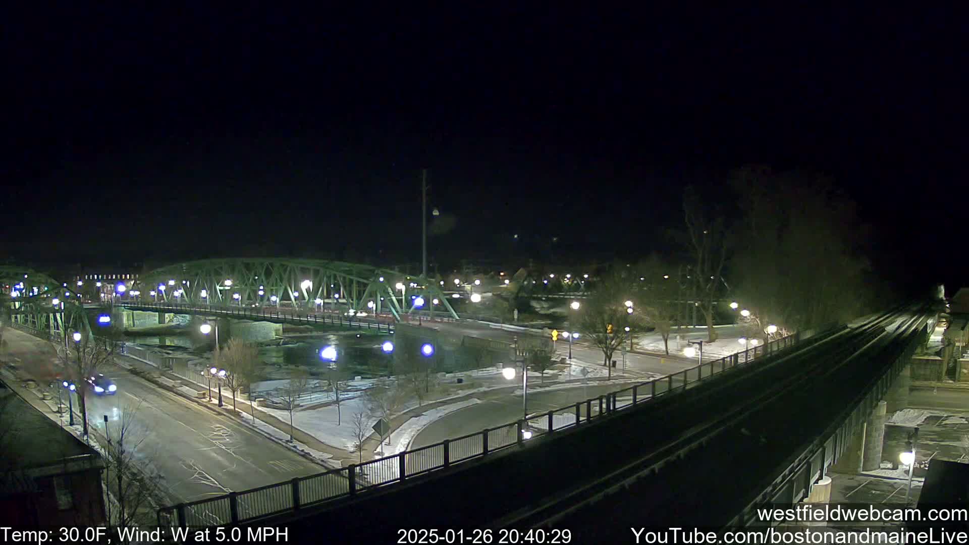 A nighttime view of a bridge over a river, with a road and train tracks intersecting, under a clear sky and a 30°F temperature with a 5 mph westerly wind.