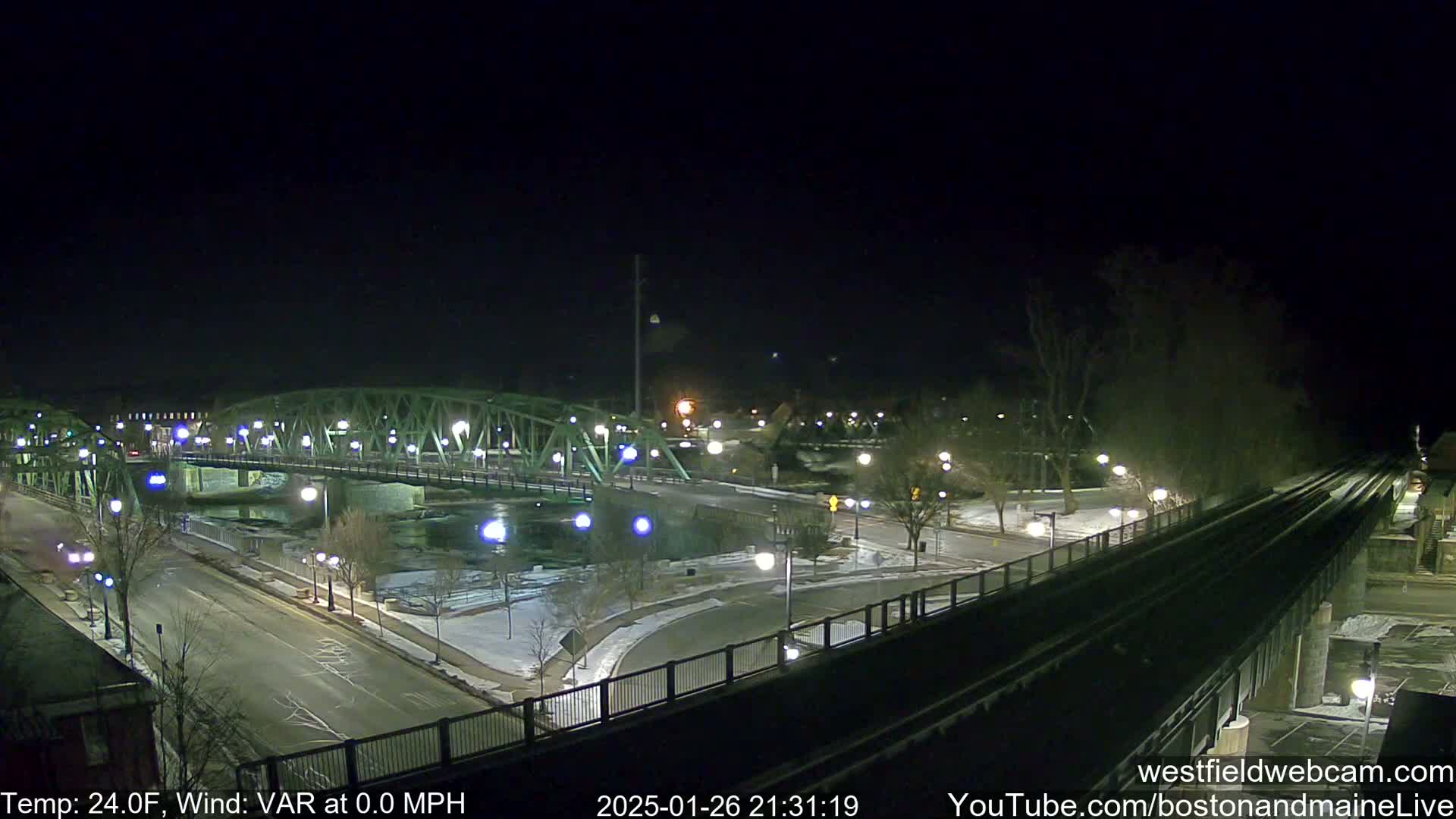 A nighttime view of a lit-up bridge over a river, with snow on the ground and a train track nearby, under a clear, dark sky.