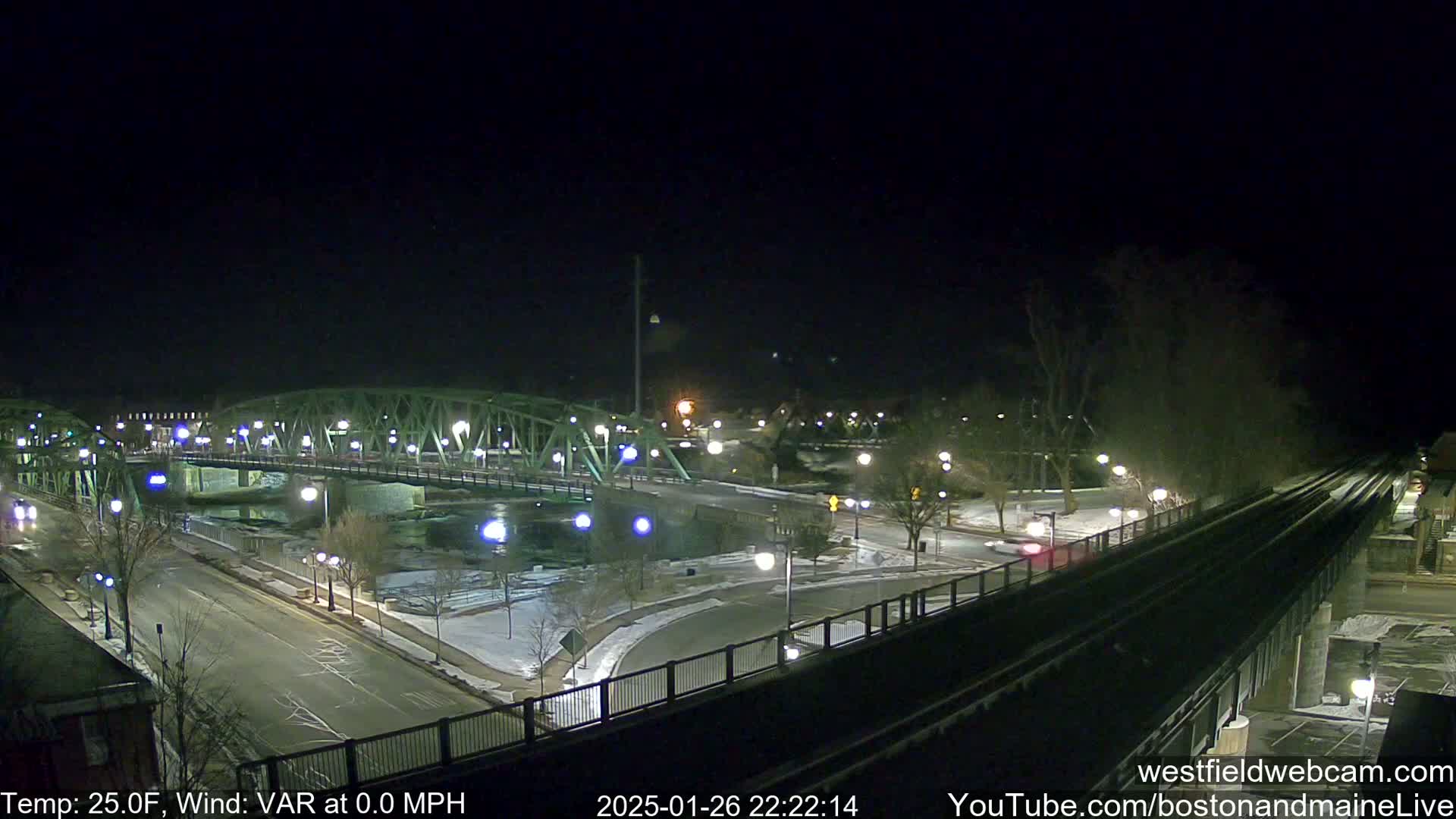 A nighttime view of a green metal bridge over a river, with snow on the ground and a 25°F temperature.