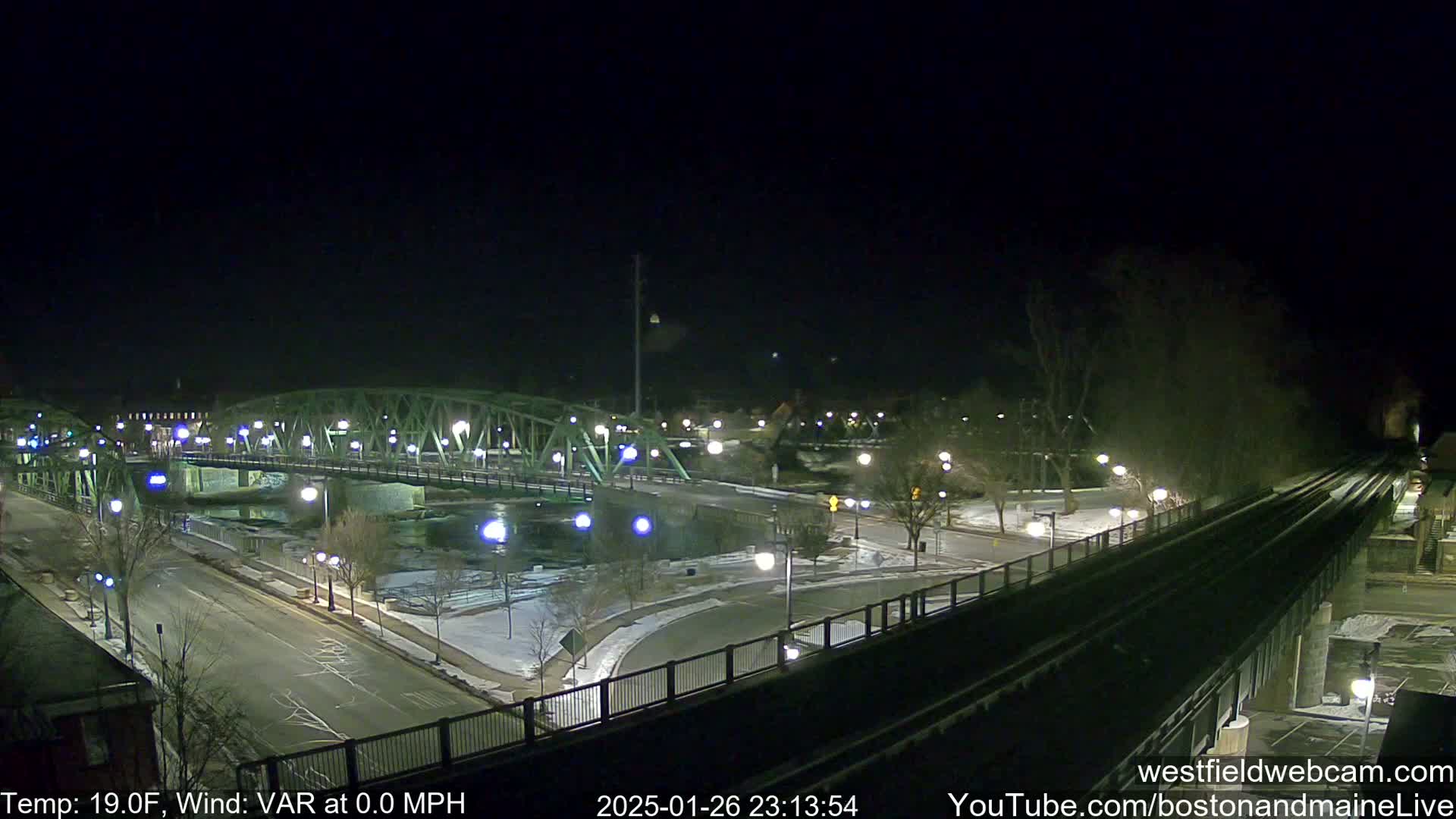 A nighttime view of a bridge over a river, with snow on the ground and a calm wind.