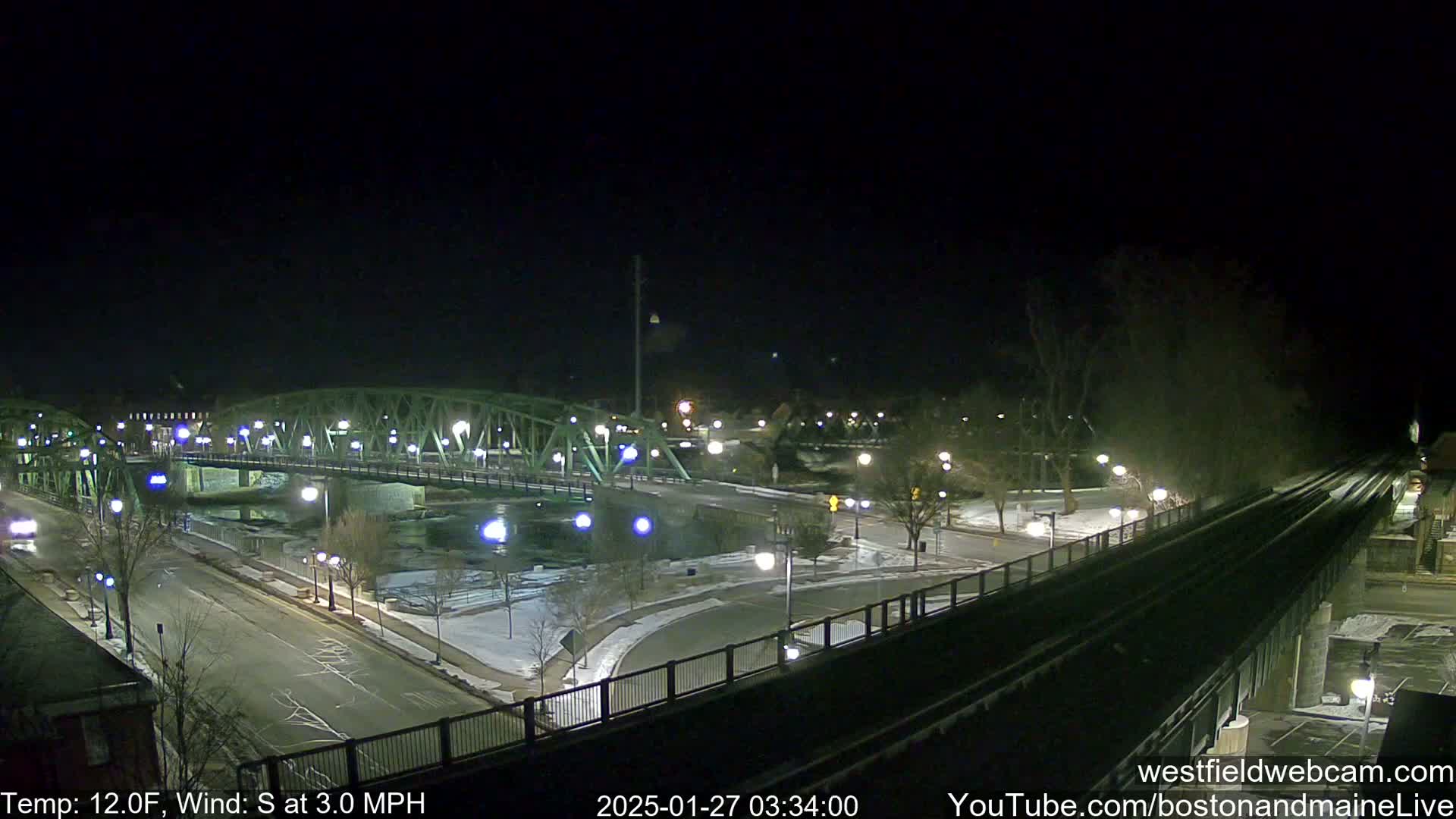 A nighttime view of a bridge over a river, with some snow on the ground and a south wind.