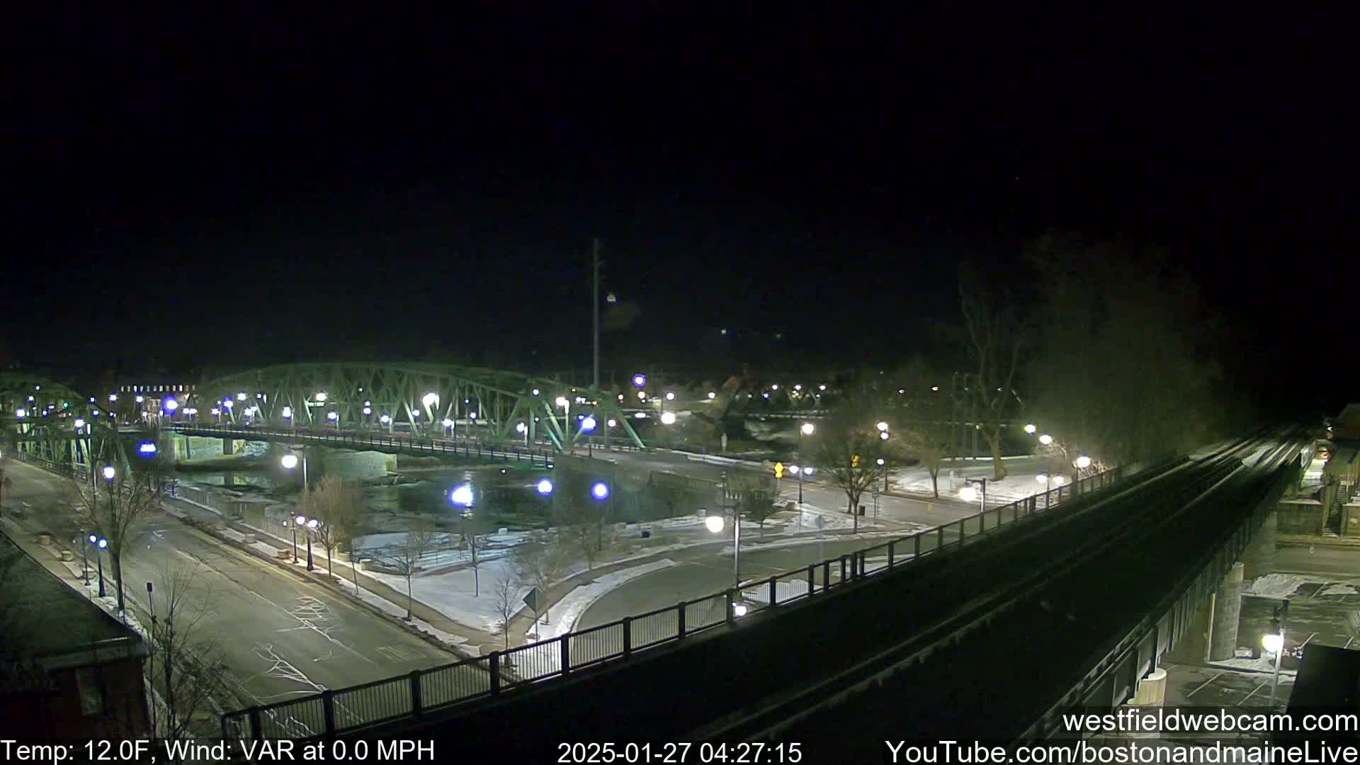 A nighttime view of a lit-up bridge spanning a river, with snow on the ground and a calm, cold wind.