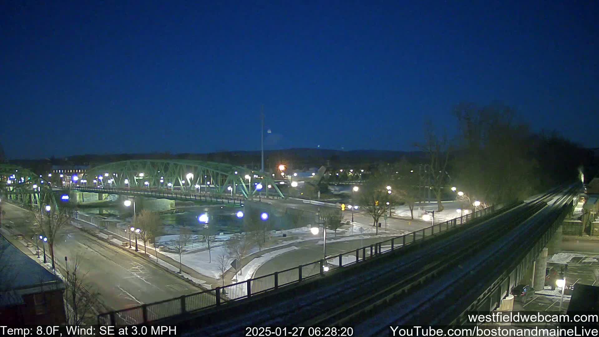 A nighttime view of a bridge over a river, with a road and train tracks in the foreground, under a clear, dark sky with light snow on the ground.