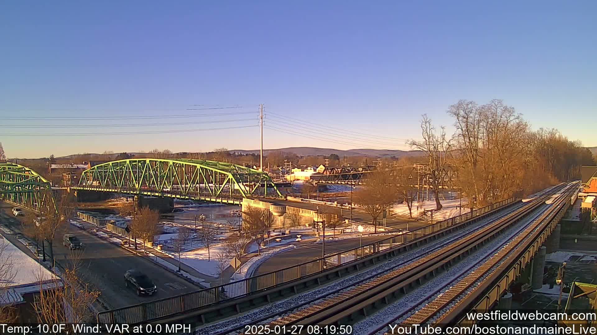 A bright, sunny day with a partly snow-covered landscape shows a green bridge over a river, with train tracks in the foreground.