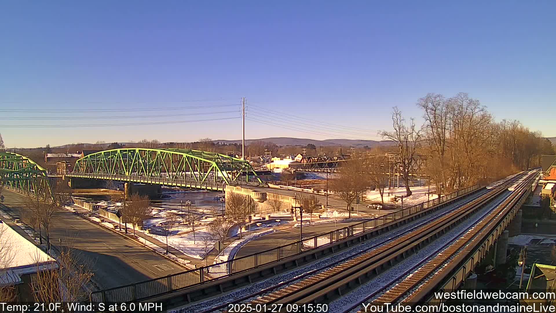 A green bridge spans a river under a clear blue sky, with train tracks and a partially snow-covered park visible in the foreground, on a cold, sunny day with a south wind.
