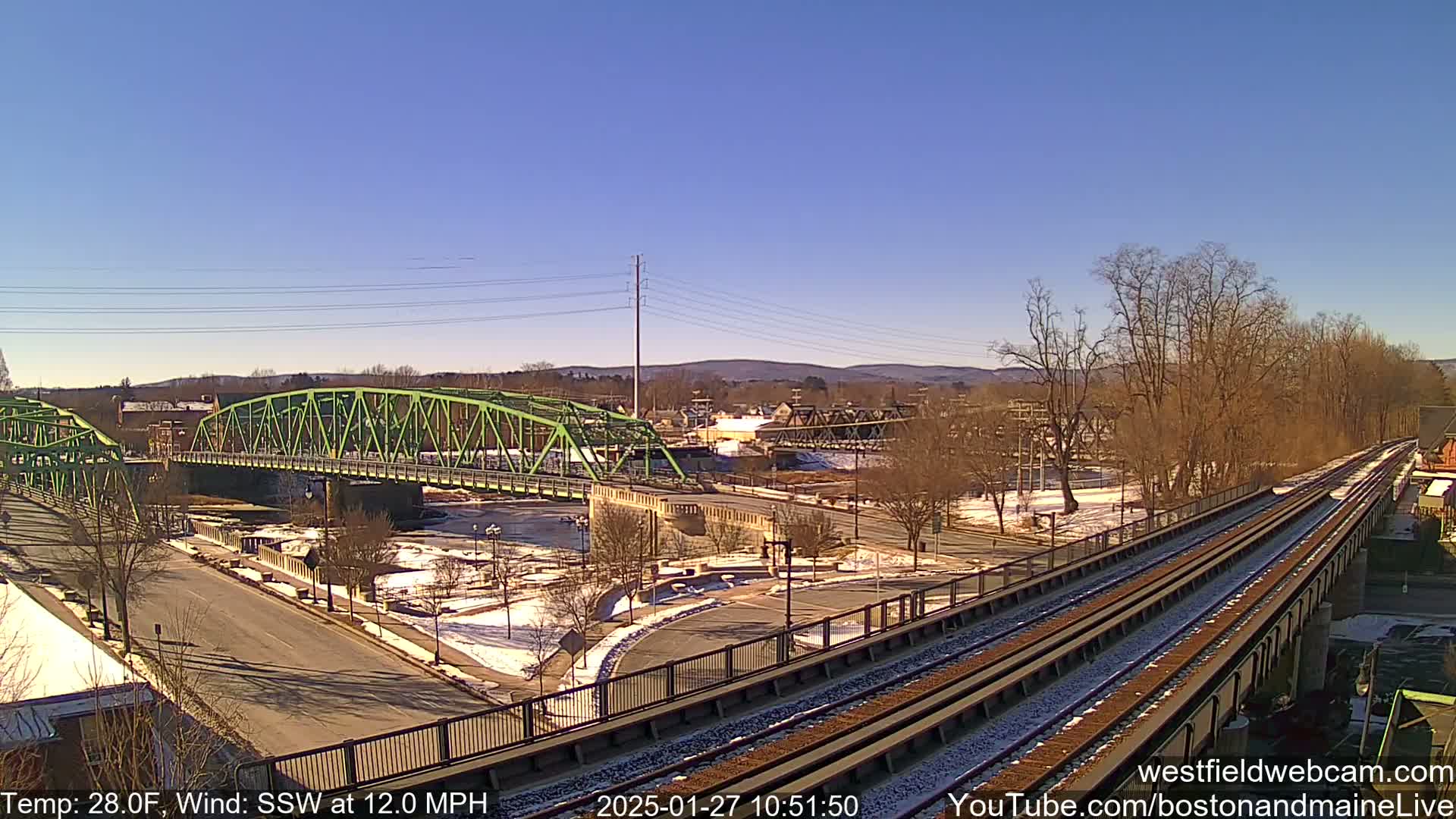 A view of a green bridge over a river, with train tracks and a town in the background on a sunny, cold day with some snow on the ground.