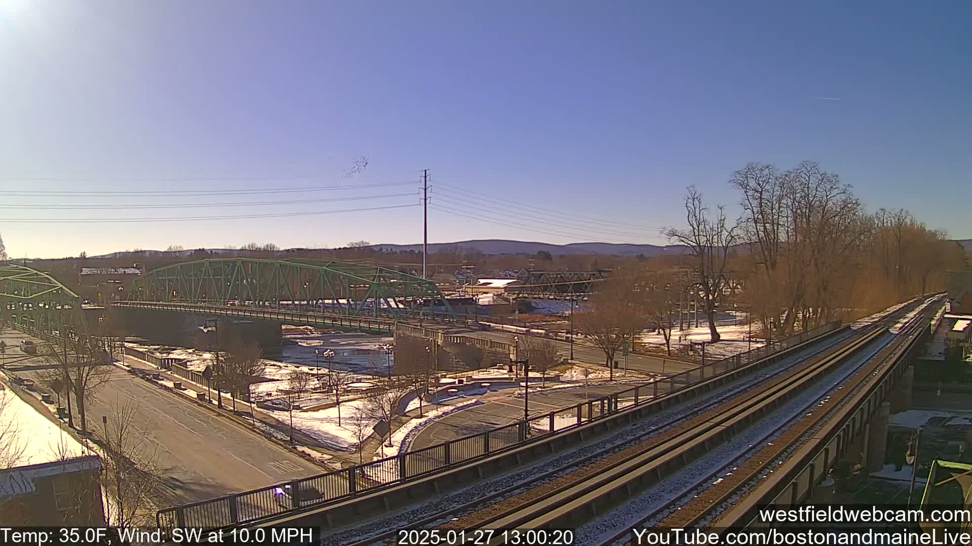 A view of two bridges crossing a river, a train track, and a town under a clear, sunny sky with a southwest wind at 10 mph and a temperature of 35°F.