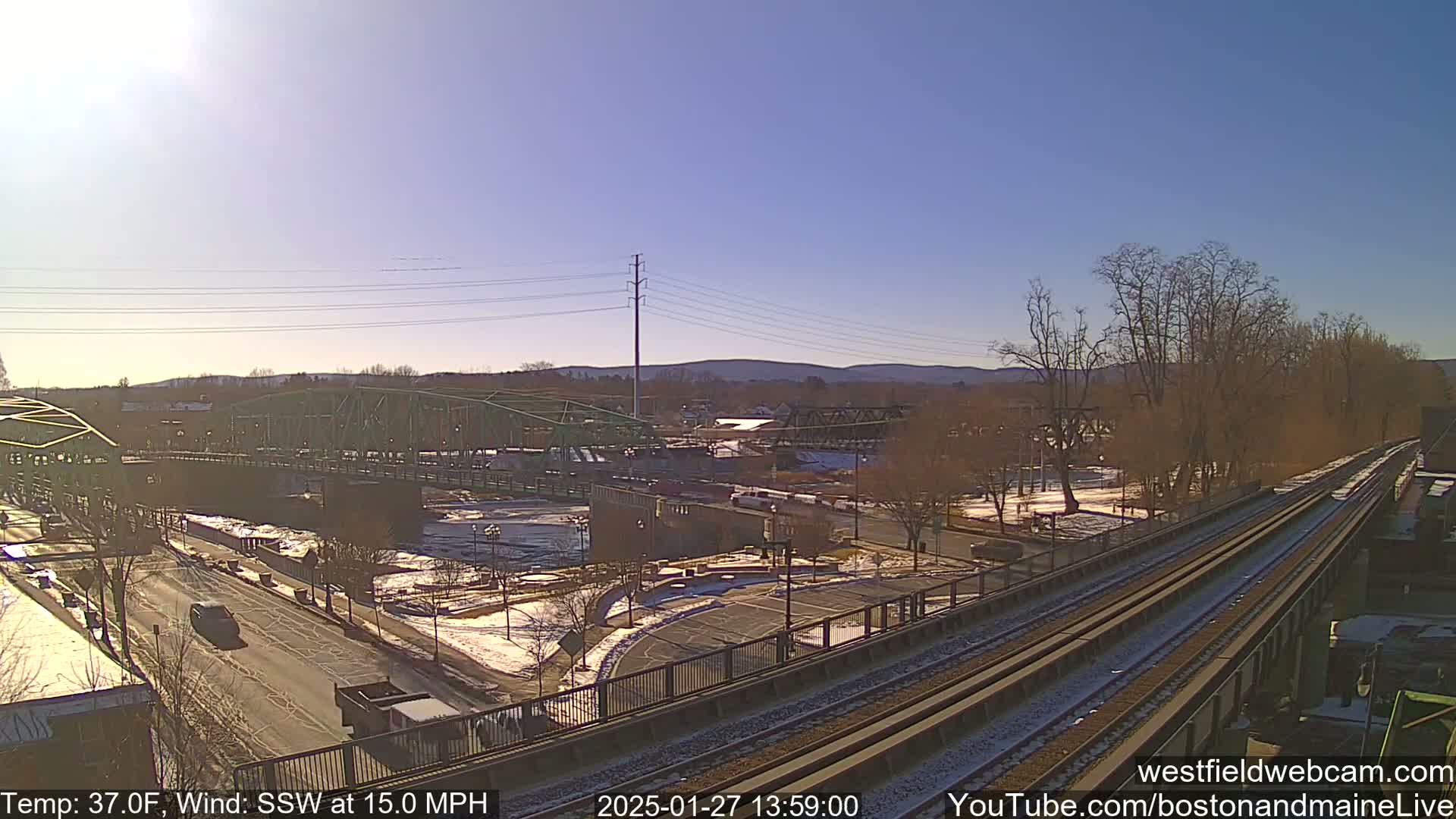 A sunny day with a southwest wind at 15 mph shows a view of a river, two bridges, a roadway, train tracks, and a sparsely populated area with some snow on the ground.