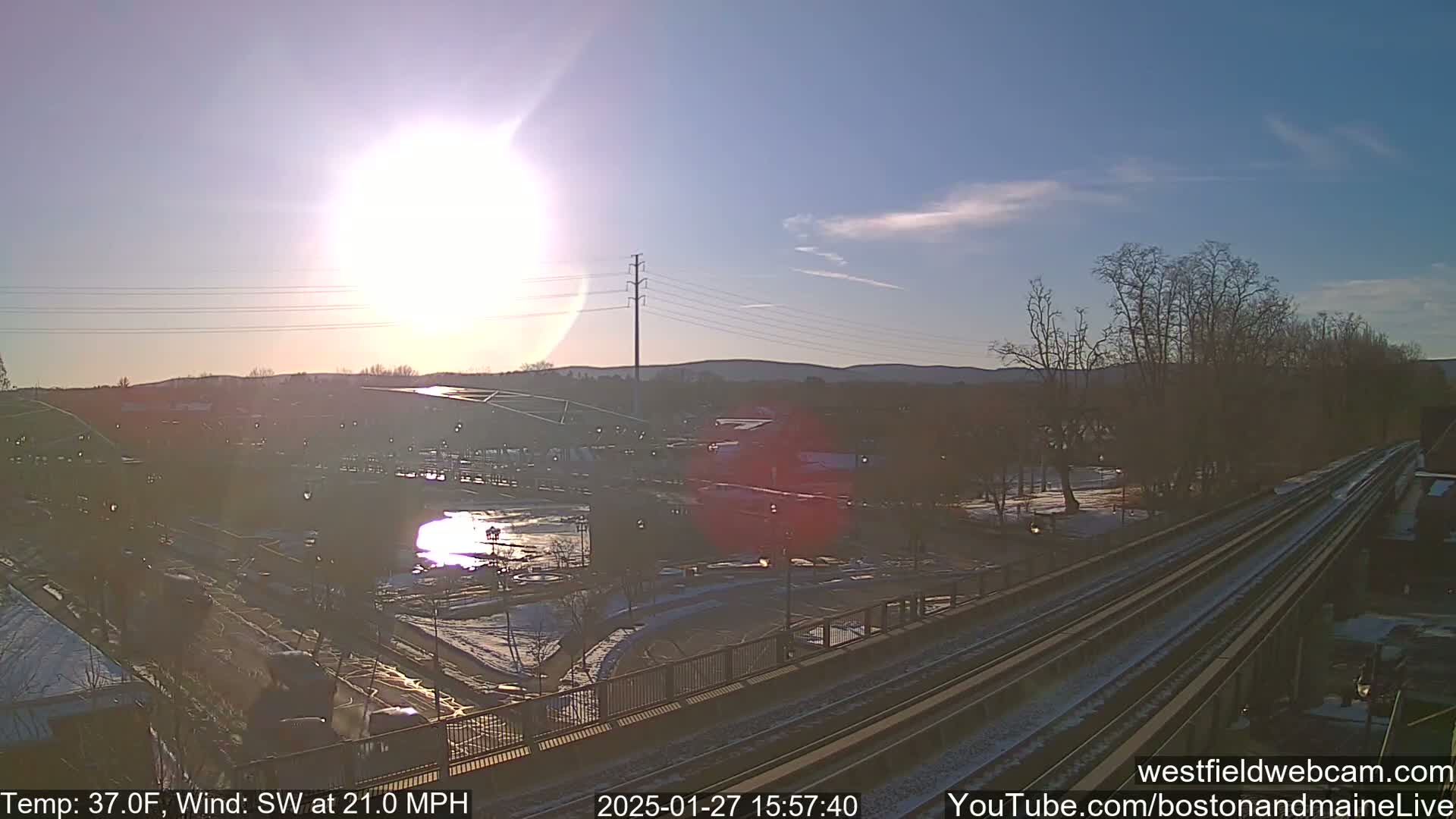 A sunny, but windy, winter scene shows train tracks, a partly snow-covered town with a reflective body of water, and bare trees.