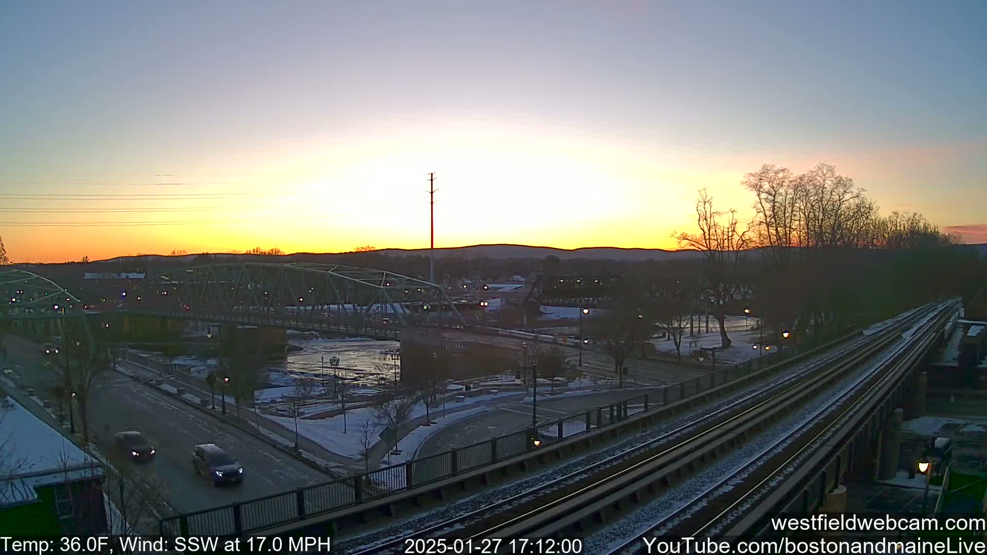 A sunset over a river with a bridge and train tracks, with some snow on the ground and a 36°F temperature and 17 mph south-southwesterly wind.