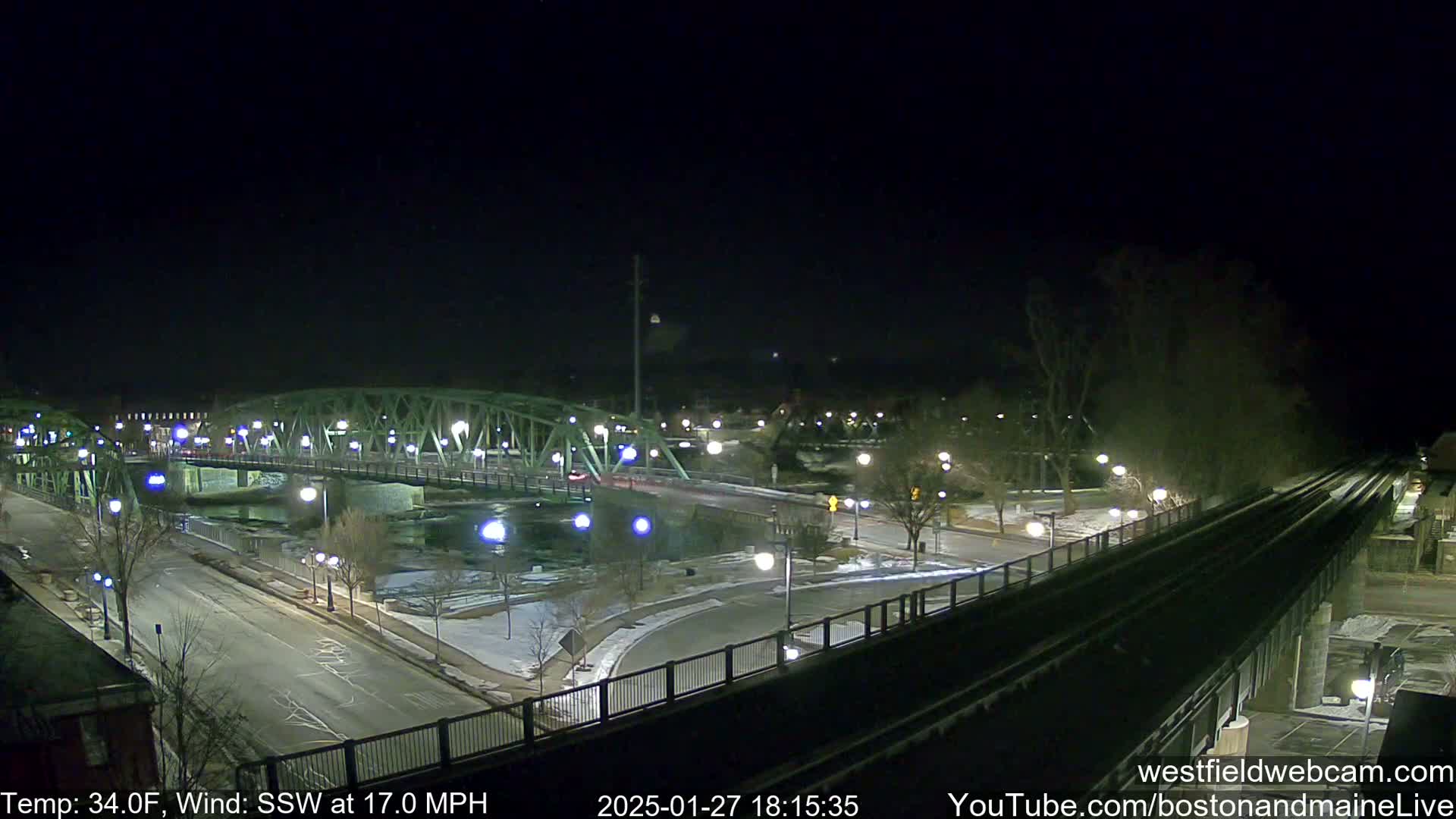 A nighttime view of a bridge over a river, with a road and train tracks intersecting nearby, under a clear sky and with a strong south-southwesterly wind.