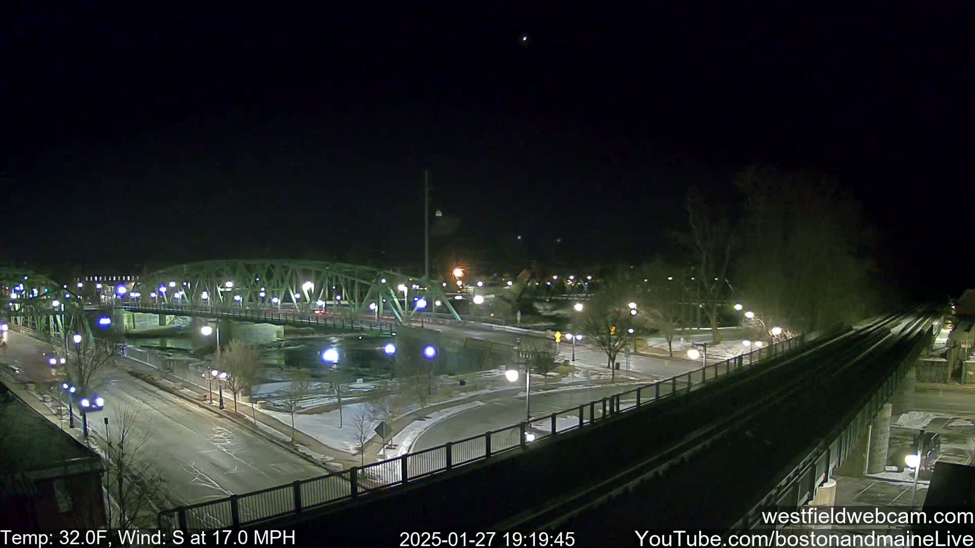 A nighttime view of a lit-up bridge over a river, with a road and train tracks visible, under a clear, dark sky with a 32°F temperature and a 17 mph south wind.