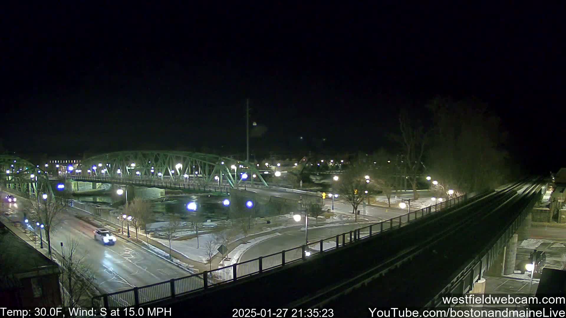 A nighttime view of a lit-up bridge spanning a river, with roads and train tracks visible, under a clear, cold, and windy sky.