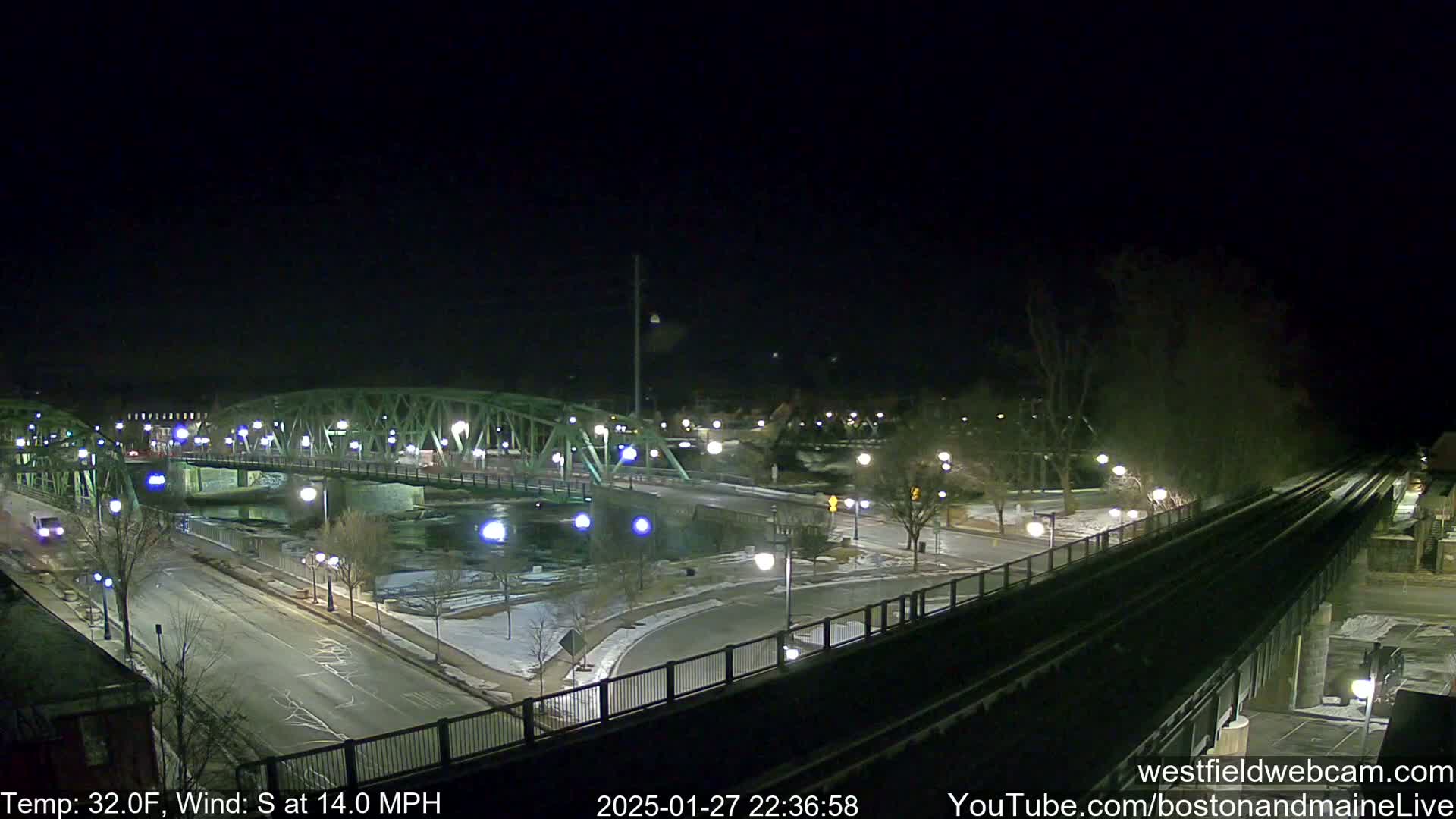 A nighttime view of a bridge over a river, with lightly snow-covered roads and surrounding areas under a clear, dark sky, a 32°F temperature and a 14 mph south wind.