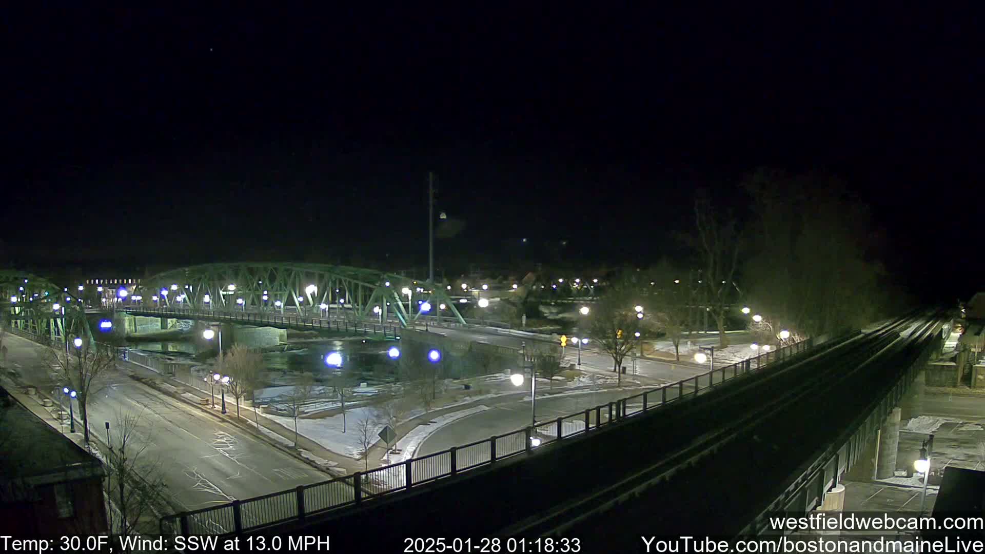A nighttime view of a green metal bridge over a river, with a road and train tracks visible, under a clear, dark sky with a south-southwesterly wind at 13 mph and a temperature of 30°F.